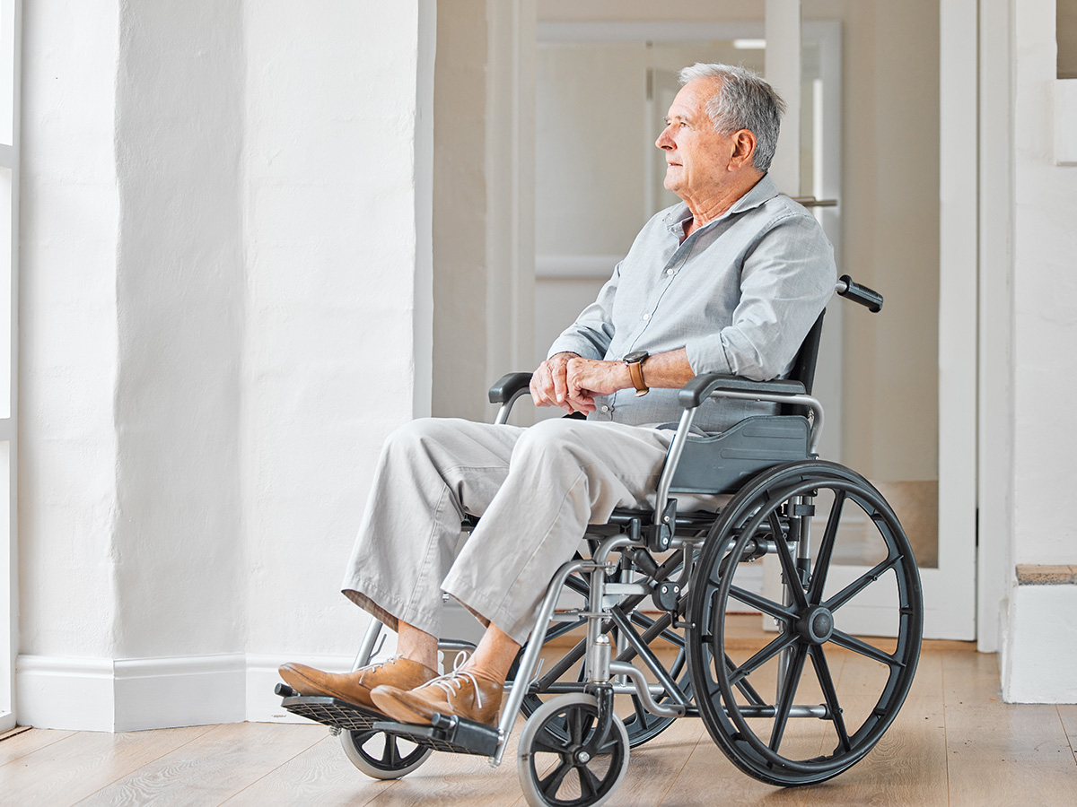 Older man seated in a wheelchair in a bright residence hallway with calm, comfortable surroundings.