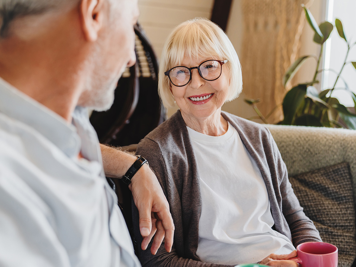 Older woman smiling while seated on a sofa with a coffee mug, chatting comfortably in a bright living space.