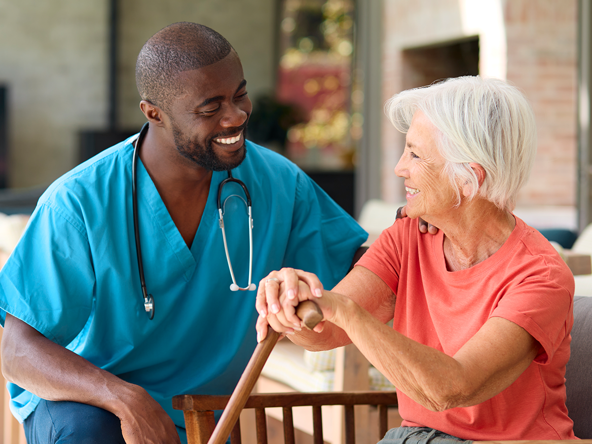 Caregiver in blue scrubs smiling with an older resident in a bright, welcoming assisted living setting.
