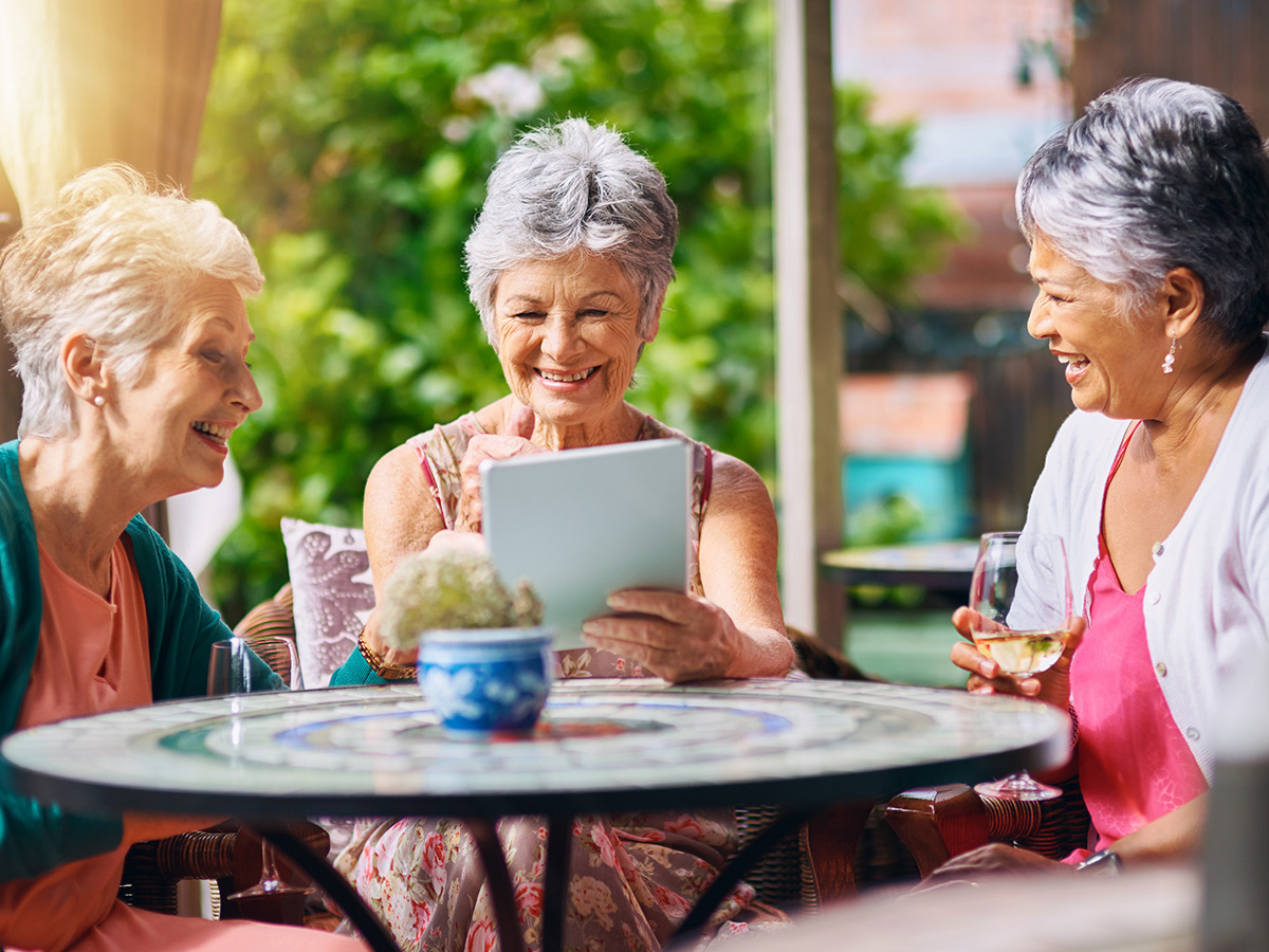 Three women enjoying conversation together on an outdoor patio table in a bright garden setting.
